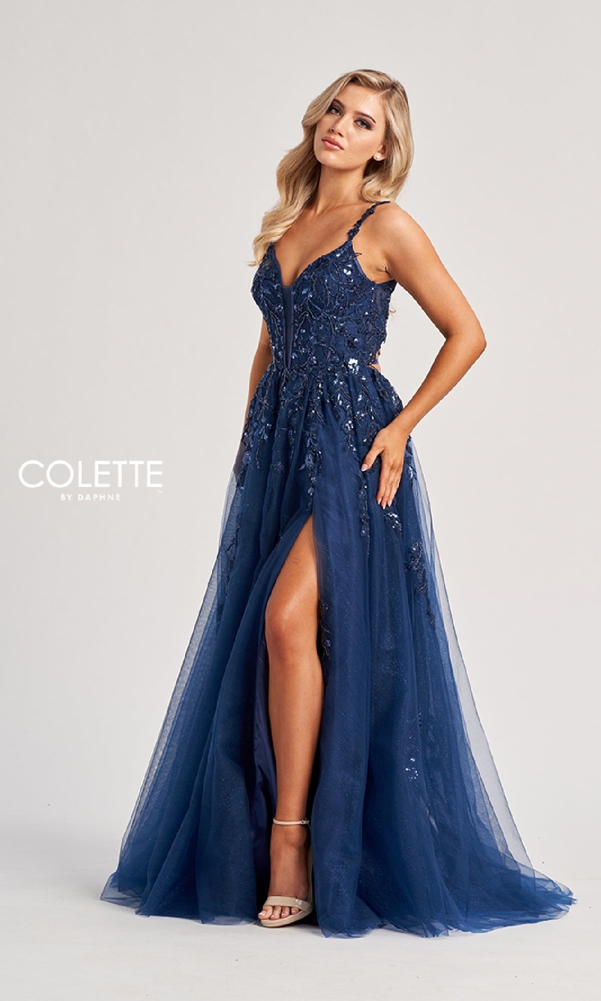 Woman wearing the Colette CL8060 Long Prom Dress, a strappy deep blue gown with sequin details and a thigh-high slit, standing against a plain light background.